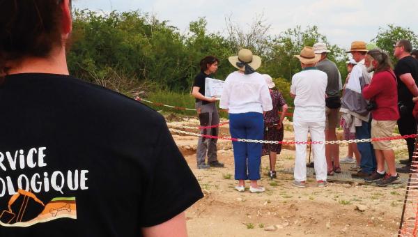 Une archéologue réalise une visite guidée de la fouille devant un groupe attentif.