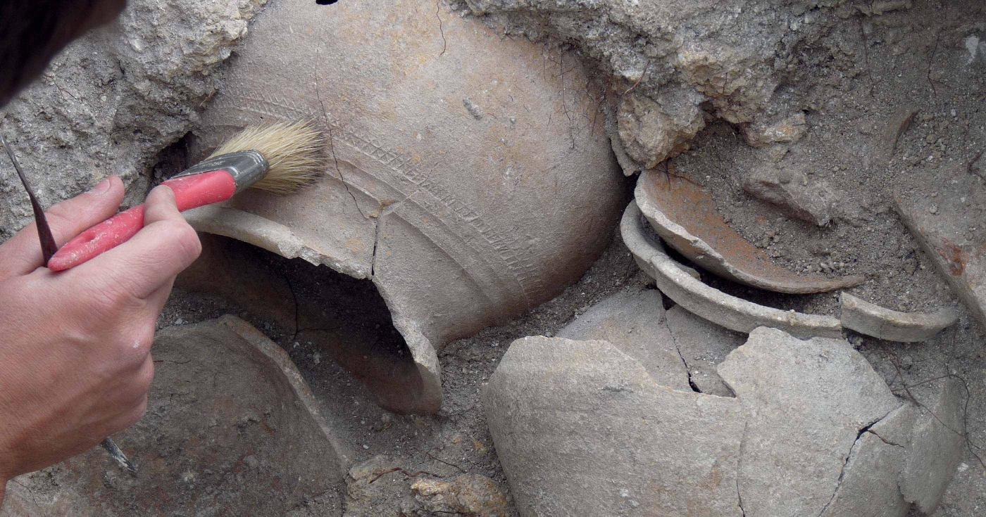 Archéologue dégageant au pinceau des céramiques enchevêtrées dans le sédiment.