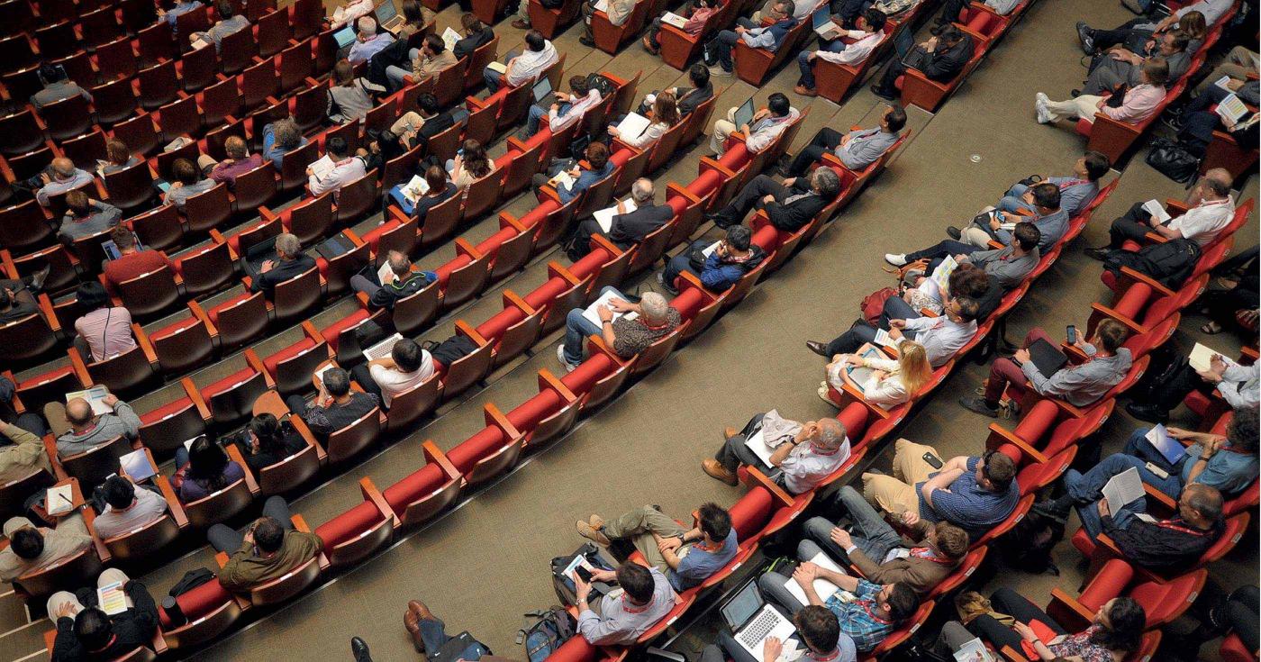 Des spectateurs sont installés sur des rangées de sièges et semblent écouter une conférence.