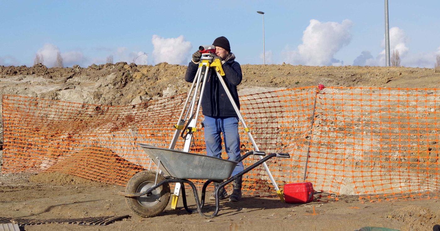 Une archéologue utilise une lunette de chantier pour prendre des mesures topographiques sur le terrain.