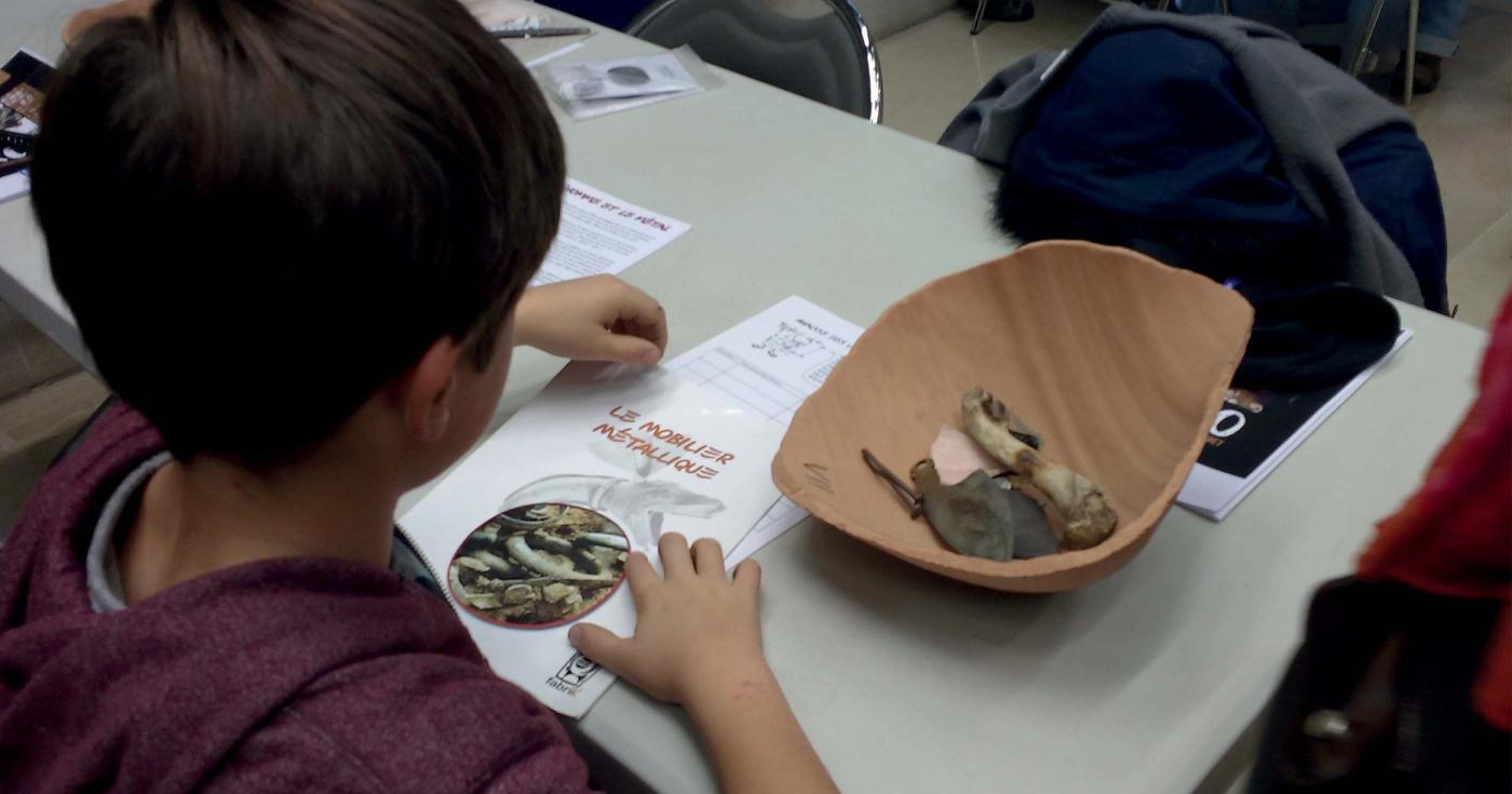 Un enfant étudie des reproductions d'objets archéologiques avec l'aide d'un livret référentiel.