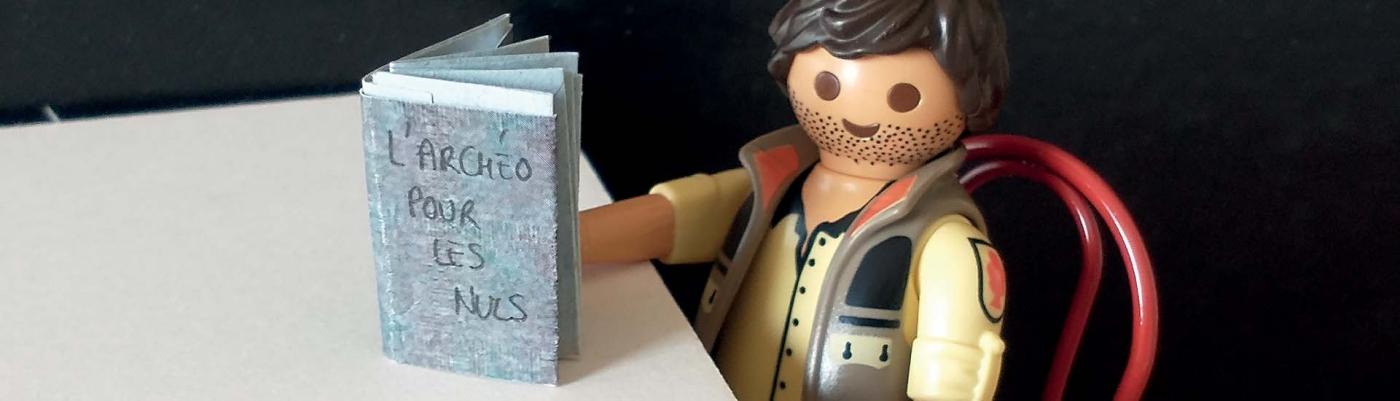Un Playmobil d'archéologue tient un livre intitulé "L'archéo pour les nuls".