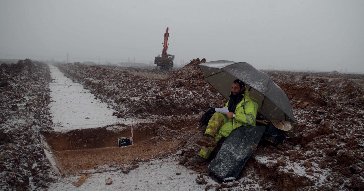 Un archéologue réalise l'enregistrement des données sur le terrain, cous la neige d'hiver, à l'abri d'un parapluie.