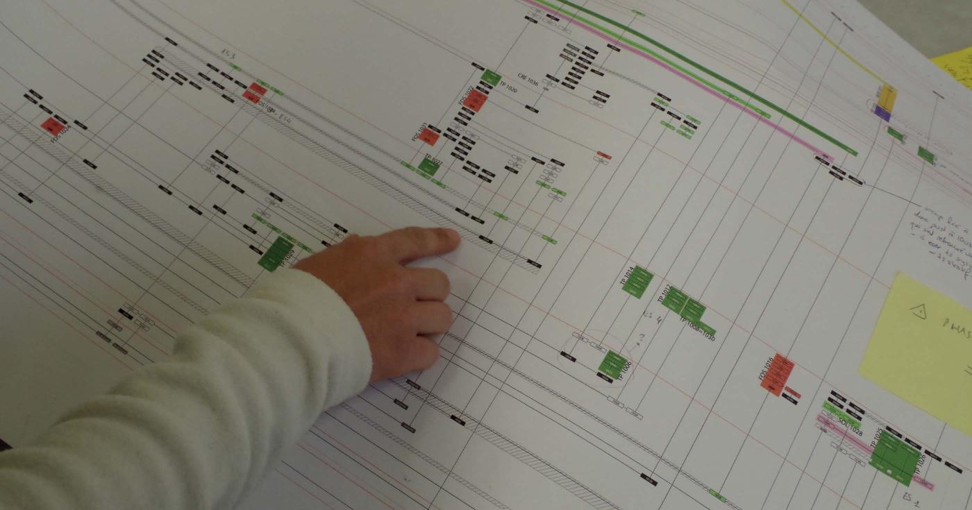 On aperçoit une main d'archéologue pointant une information sur un grand papier imprimé. Ce dernier contient une grande quantité de données reliées entre elles comme un arbre généalogique.